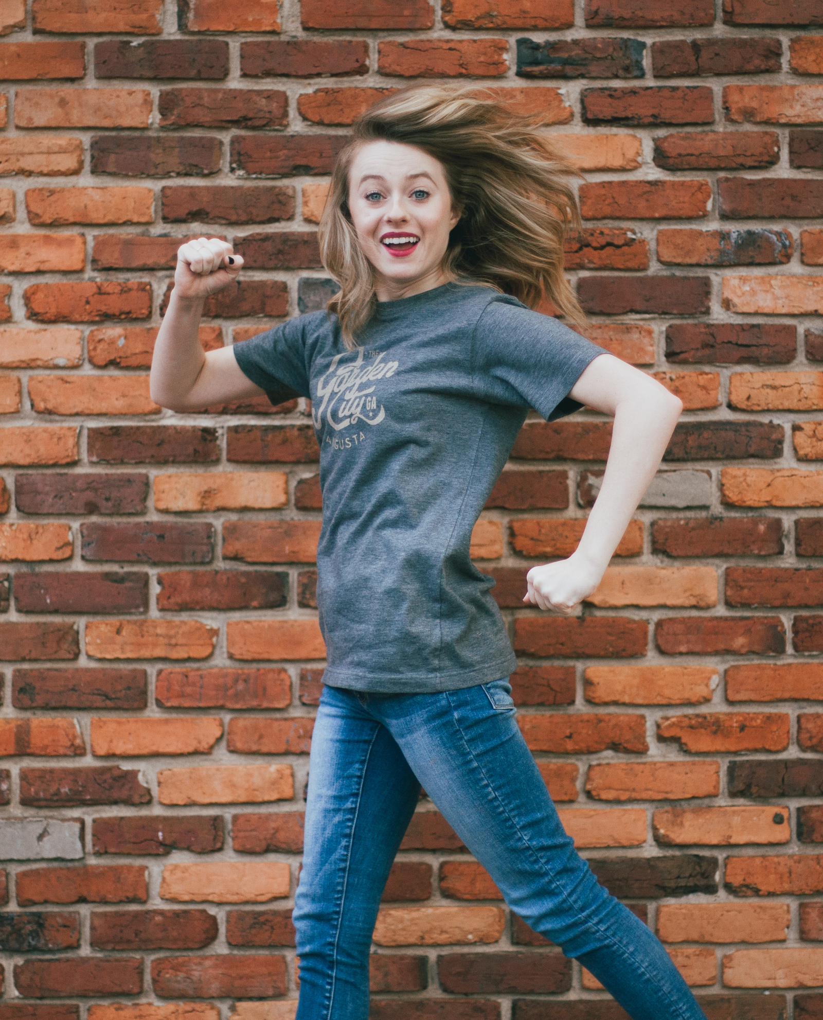 Woman jumping in front of a brick wall wearing blue vintage Garden City shirt