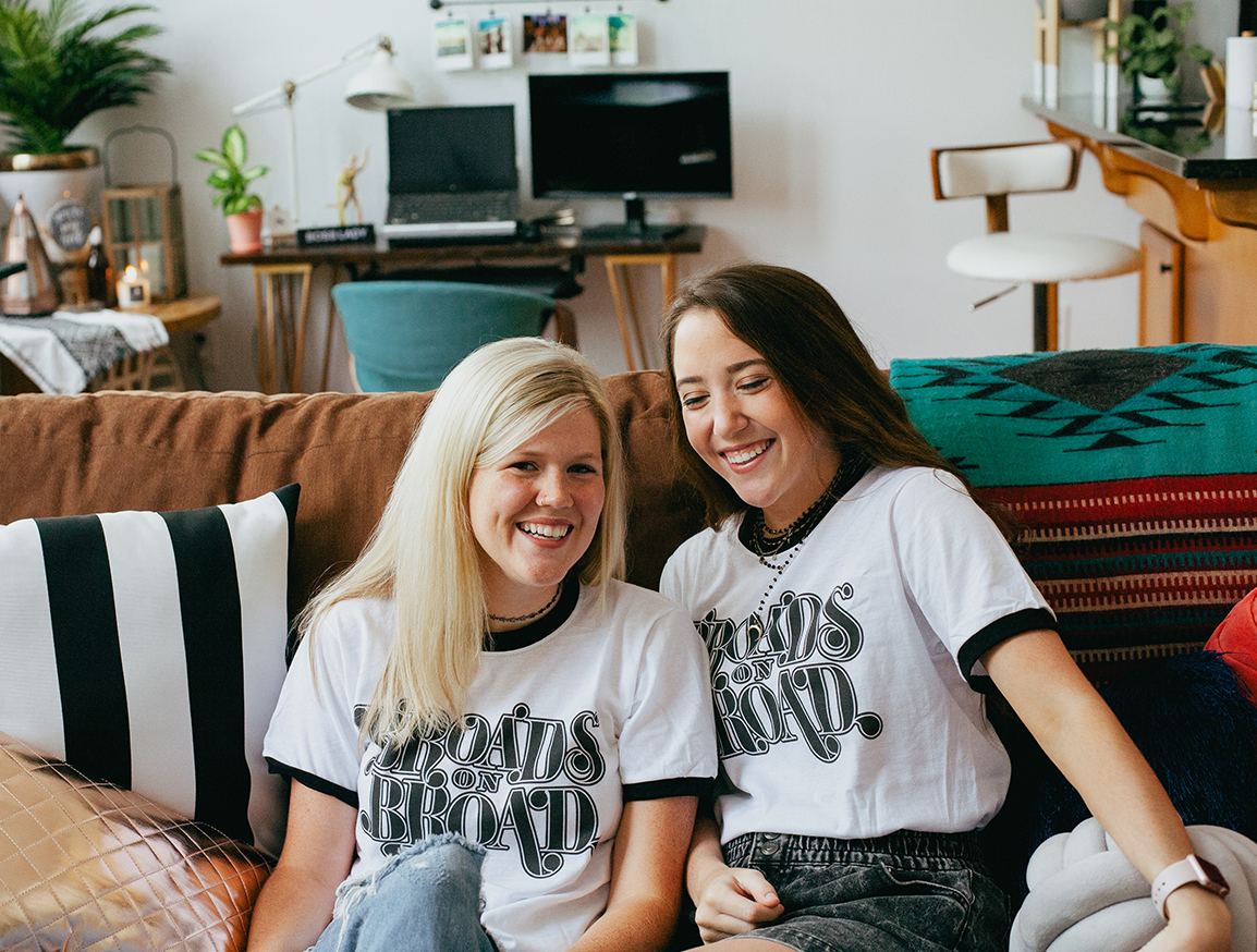 Two women sitting on a couch and wearing Broads on Broad shirts