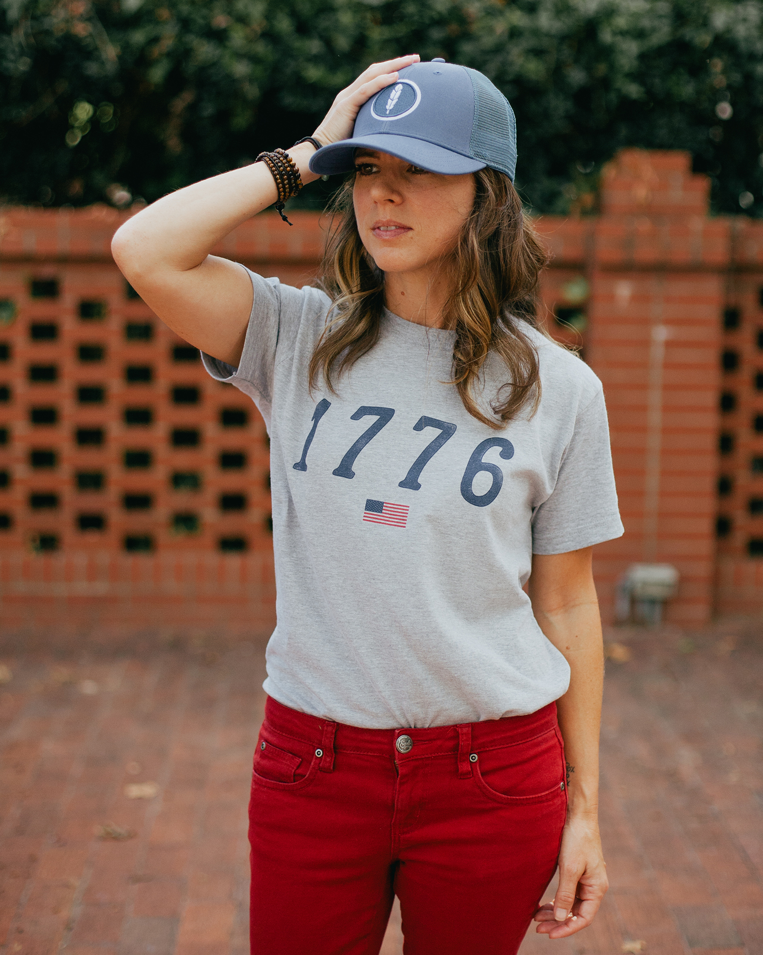 Woman wearing gray 1776 USA shirt with hand on blue feather trucker hat
