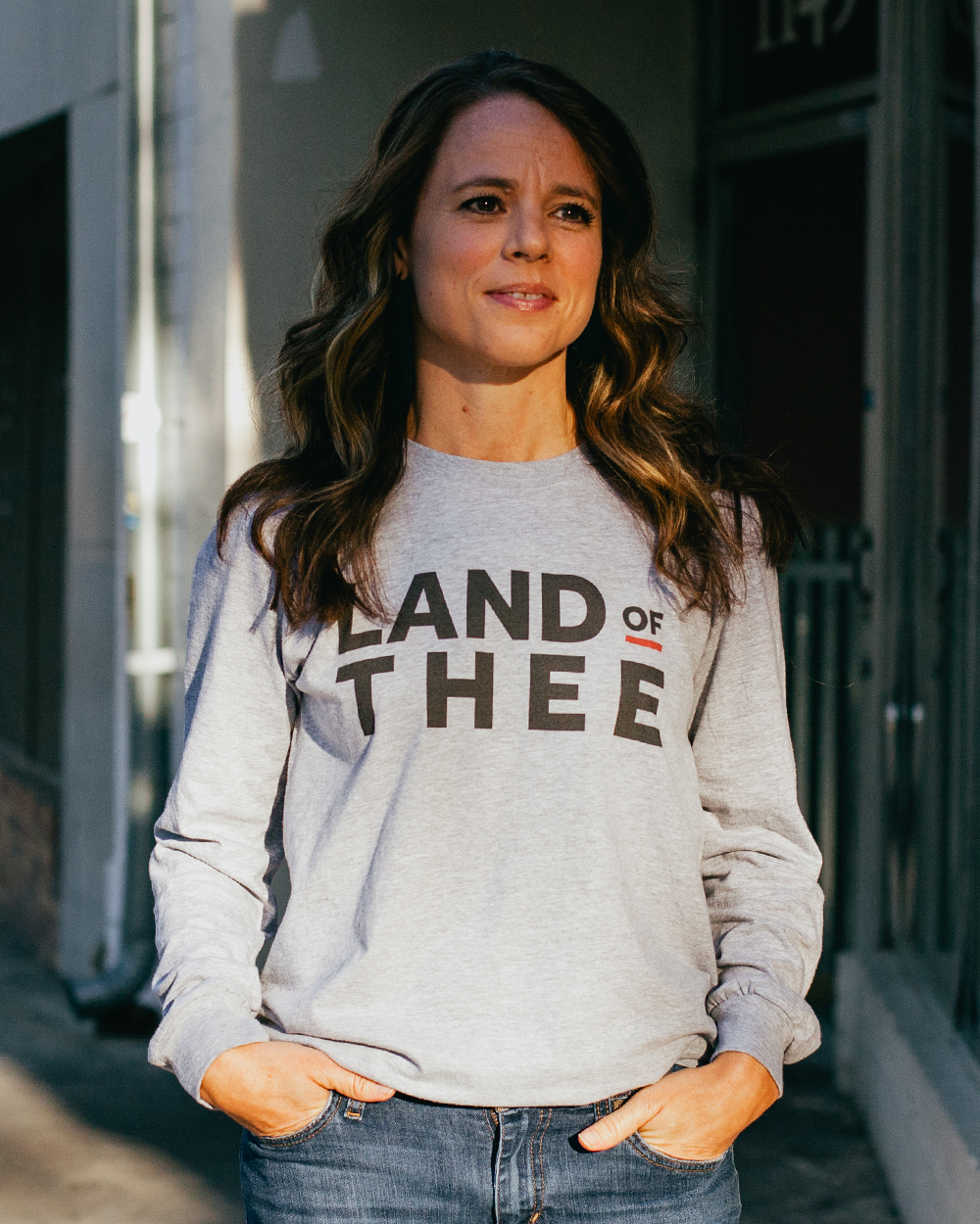 Woman wearing gray long sleeve Land of Thee shirt with jeans